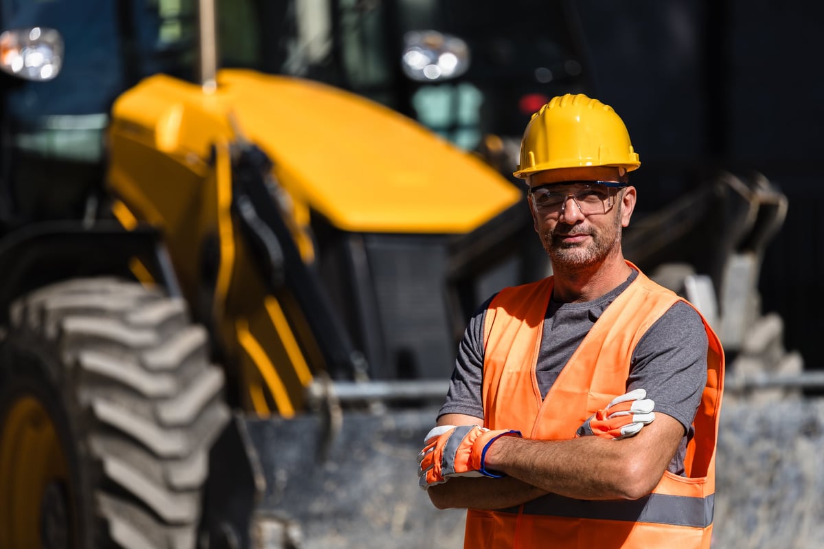 A construction worker in an orange vest and hard hat crossing arms in front of yellow machinery